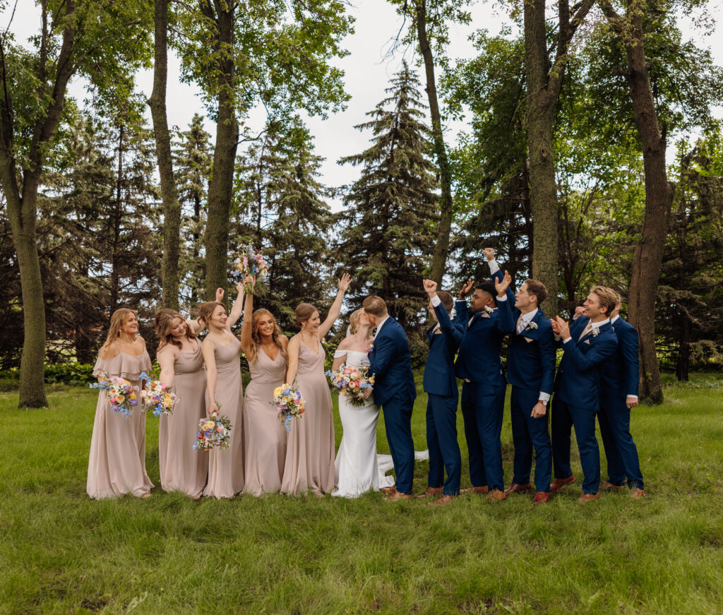 bride and groom share a kiss outside surrounded by pine trees as their bridal party wearing navy blue suits and blush pink dresses cheers with their arms in the air in celebration