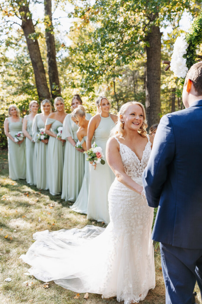 Bride holding hands with her groom during an outdoor Minnesota fall ceremony as her bridesmaids in soft teal dresses stand smiling behind her