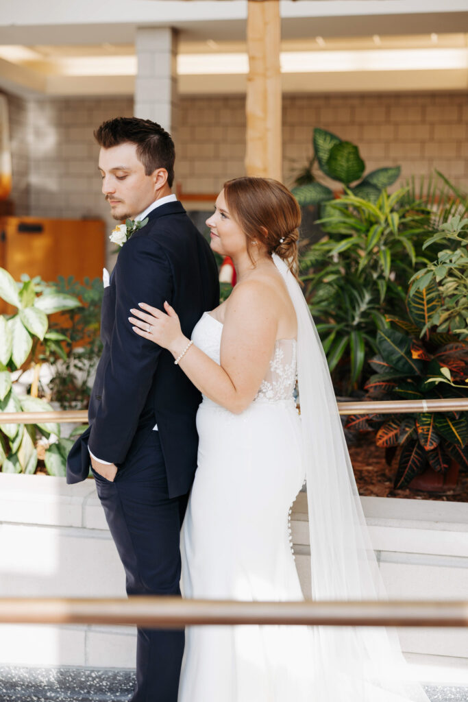 bride stands behind her groom with her left hand gently on his bicep smiling as he looks softly over his shoulder at her