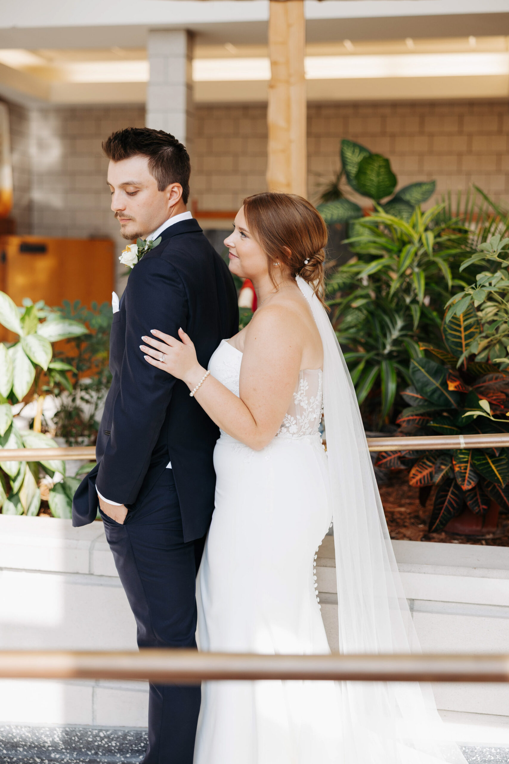 bride stands behind her groom with her left hand gently on his bicep smiling as he looks softly over his shoulder at her