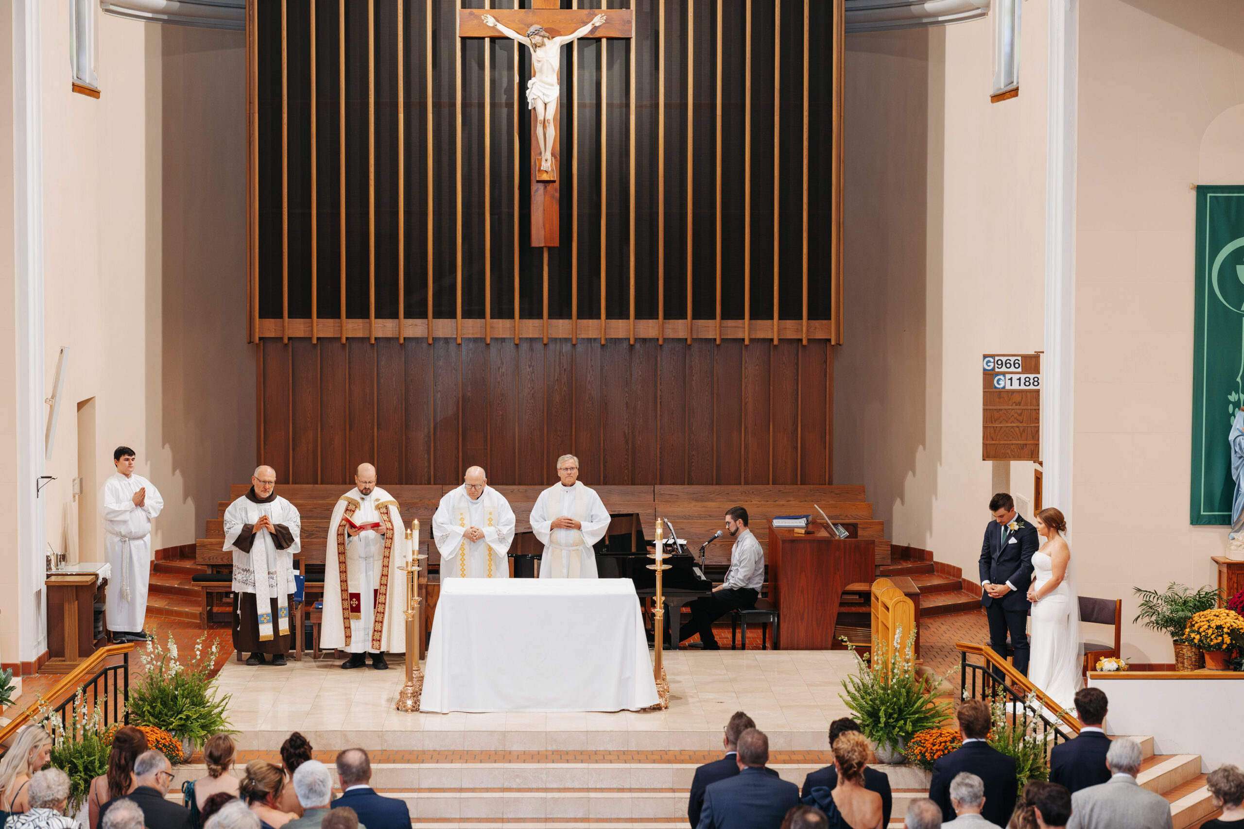 beautiful catholic church indoor ceremony including 4 priests standing at the alter with a large wooden cross at the center of the back fluted wall bride and groom stand to the right listening intently
