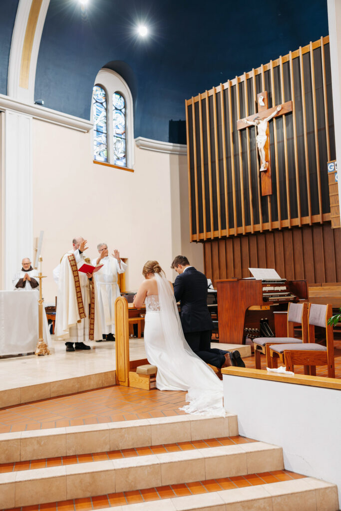 bride and groom kneel at the alter during an indoor catholic wedding ceremony as the priests lift their hands praying over them
