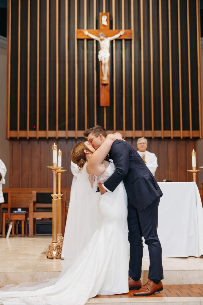 bride and groom share their first kiss at the alter of a beautifully catholic church surrounded by wood fluted walls