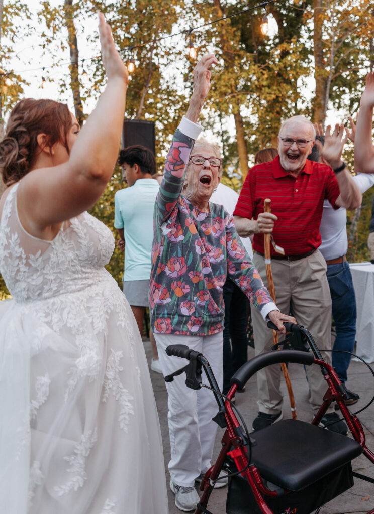 elderly women with a walker lifts her hands in the air singing and dancing with the bride on an outdoor dance floor mid summer