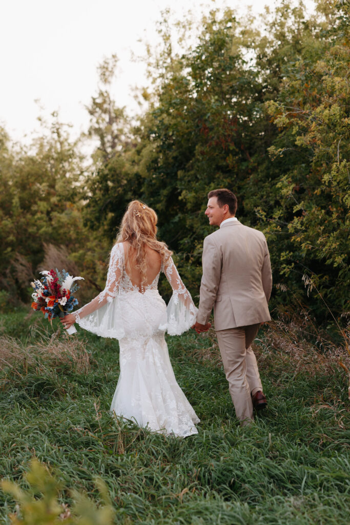 bride in a long sleeve laced bridal gown holding a bojo bouquet of flowers walks through a field of tall grass with her groom in a tan suit