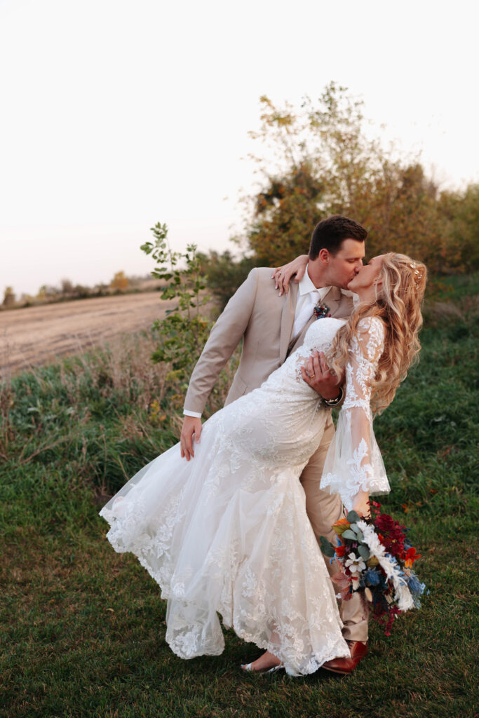 groom in a tan suit dips his bride for a kiss as she wears a lace long sleeved bridal gown during sunset hours in a field of tall grass