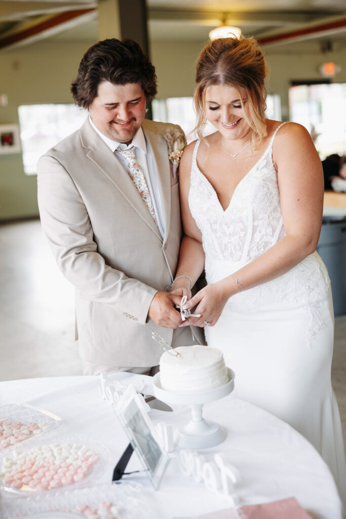 groom in a tan suit and bride in a mermaid lace gown share a moment of laughter while cutting their wedding cake at their reception