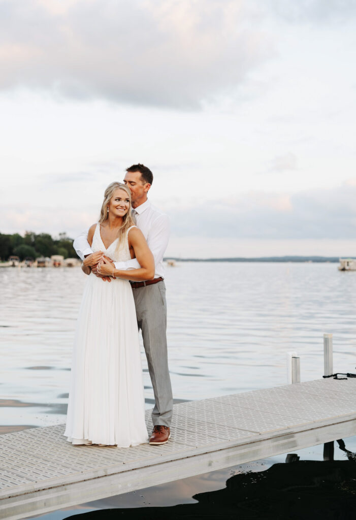 bride and groom stand on a dock overlooking the lake as the groom holds his bride wearing an A line gown from behind and kisses the back of her head as the sunset sets behind them