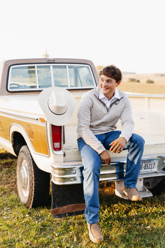 young man posed on the tailgate of an old ford pick up truck in jeans and a sweater with a cowboy hat hanging next to him