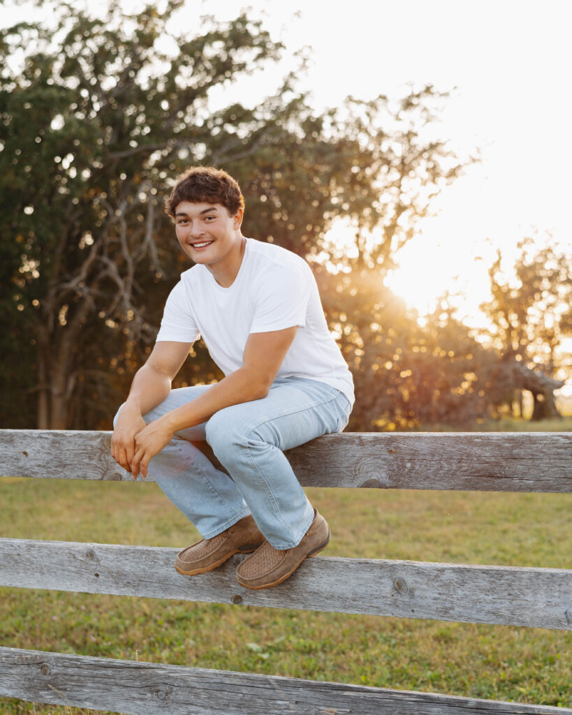 young man in blue jeans a white t shirt sitting on top of a wooden fence outside with a sunset going down behind him