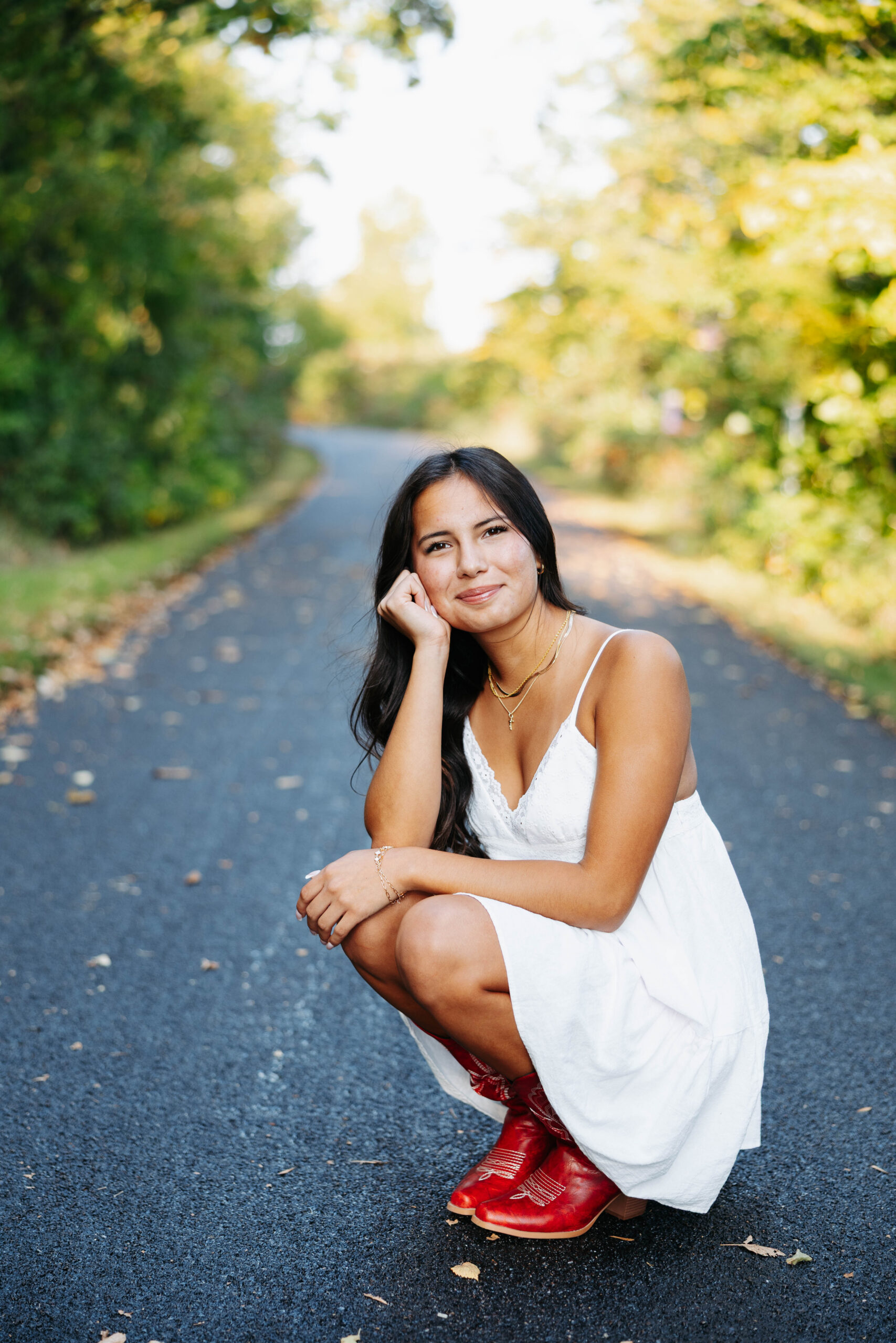 women crouched in a white summer dress wearing red cowboy boots with one hand on her chin and theo ther on her lap on a paved road through a wooded area