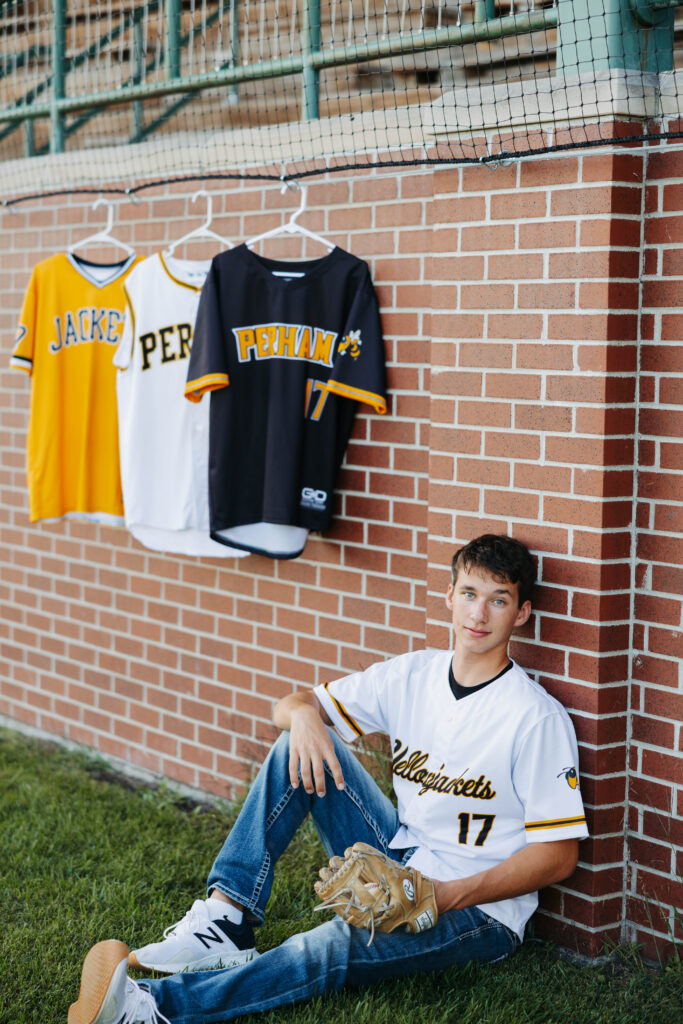 young man leaning against a brick wall in a white baseball jersey while other jerseys hang in the background and he has one hand in a baseball glove