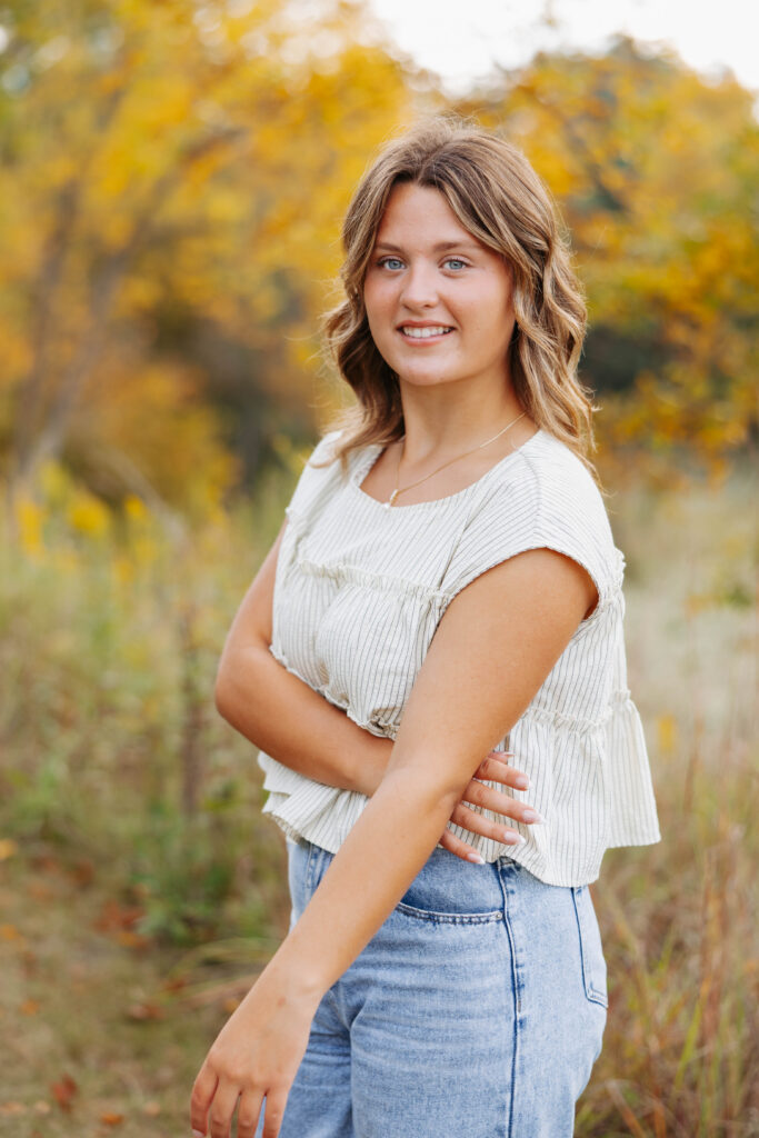 young women in jeans and a white blouse poses in a field with fall colors surrounding