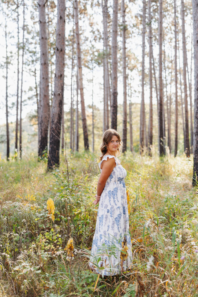 young women in a long white and blue summer dress standing looking over her shoulder outdoors surrounded by tall pine trees and grass 