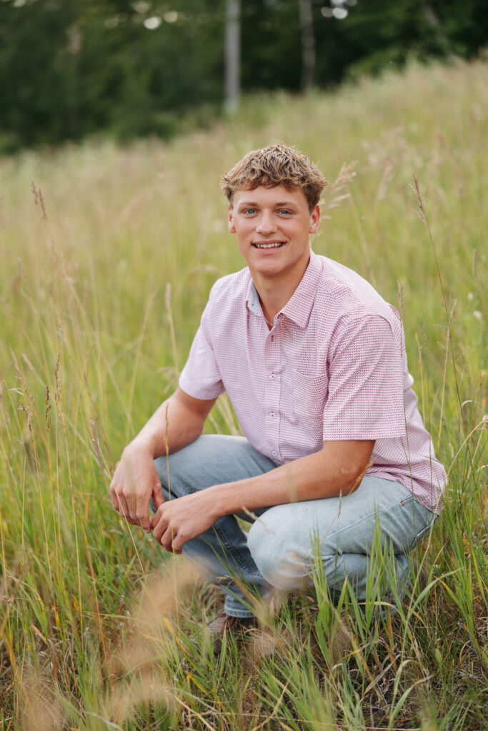 young man in jeans and a pink shirt crouches in a field of tall grass