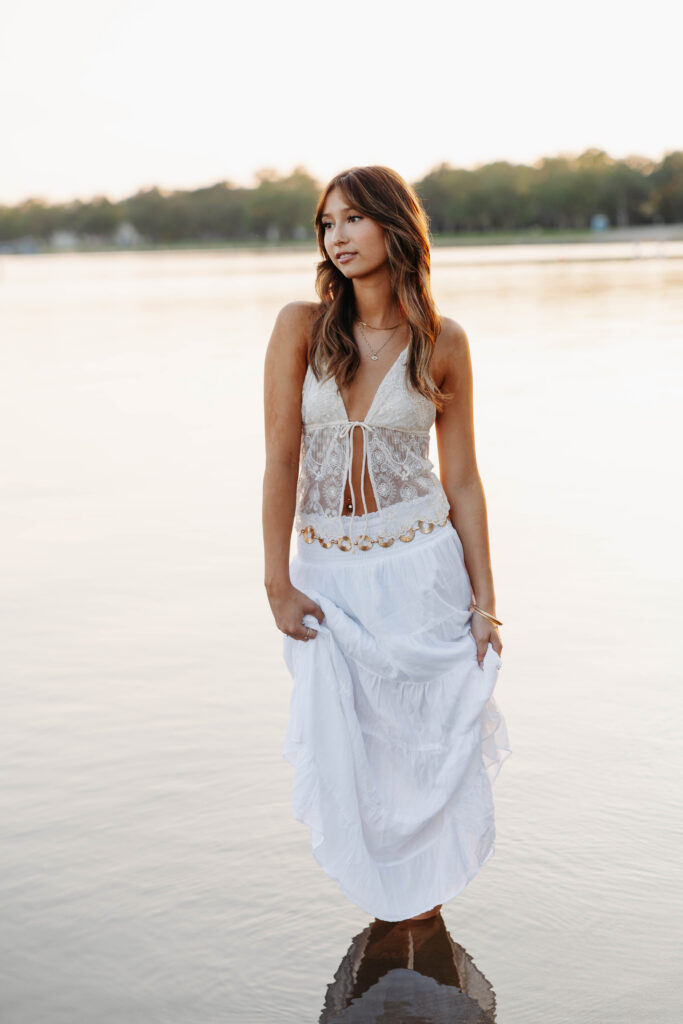 brunette young women in a long flowing skirt and a lace top walking in shallow water of a lake with the sunset behind her