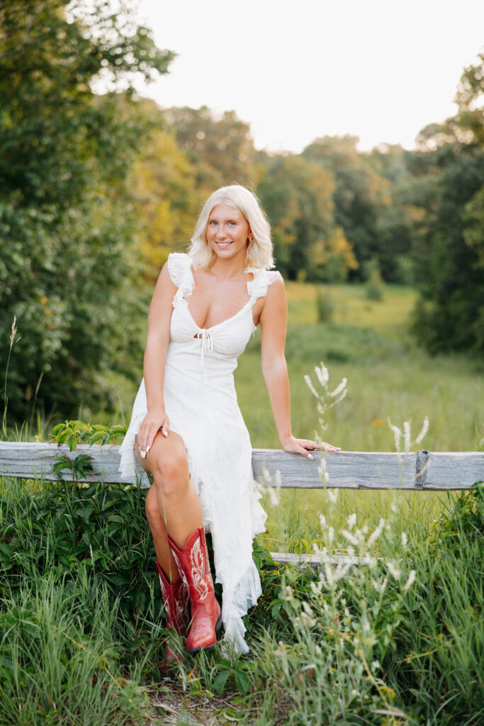 young women in a long white summer dress and red cowboy boots sitting on a wooden fence in a wooded area with tall grass