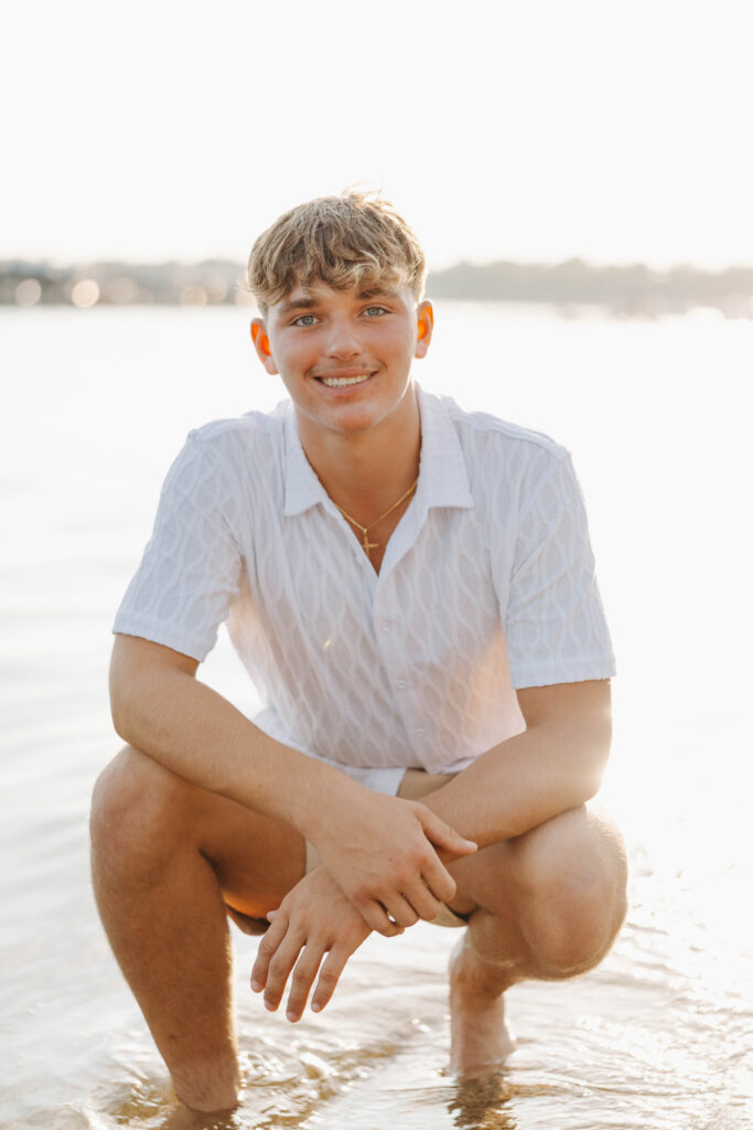 young man crouched down in shallow water in a white shirt and tan shorts smiling with the sunset behind him